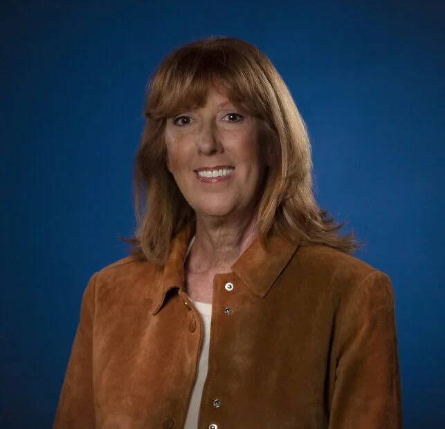 Professional headshot of a middle-aged woman with red hair wearing a brown jacket against a blue background.