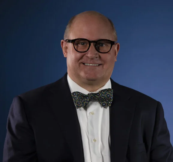 Professional portrait of a smiling bald man wearing glasses, a dark suit, white shirt, and patterned bow tie.