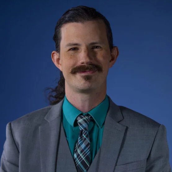 Professional headshot of a man with dark hair and mustache, wearing a gray suit and teal shirt with a patterned tie.