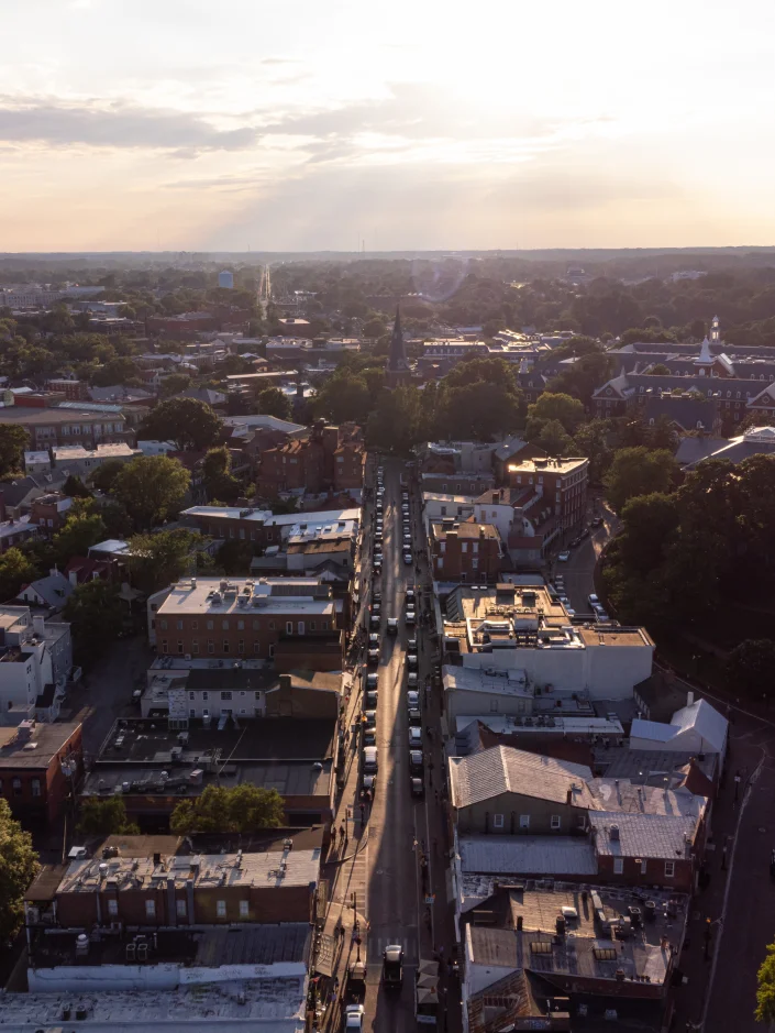 Aerial view of a city street lined with buildings and trees under a cloudy sky at sunset.
