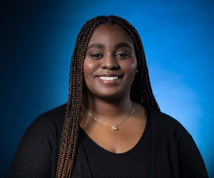 Portrait of a smiling woman with long braided hair, wearing a black top, against a blue background.
