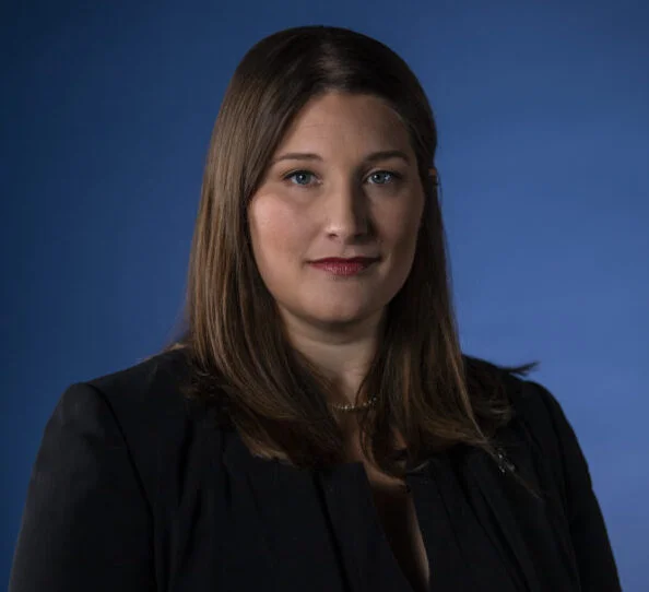 Professional portrait of a woman with brown hair, wearing a black blazer against a blue background.